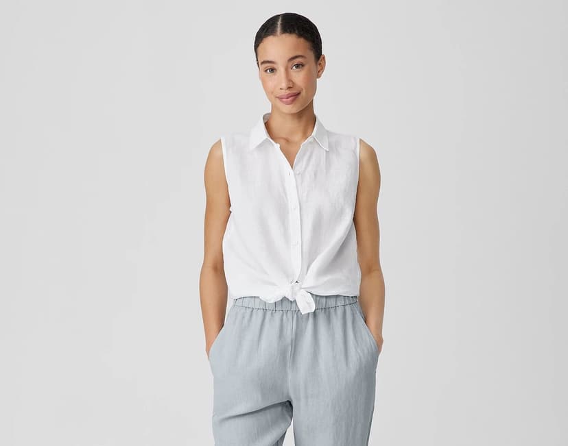 A woman with her hair pulled back wears a white, sleeveless linen blouse with a knot tied in front of the stomach and grey linen pants. She has her hands tucked in the pockets.