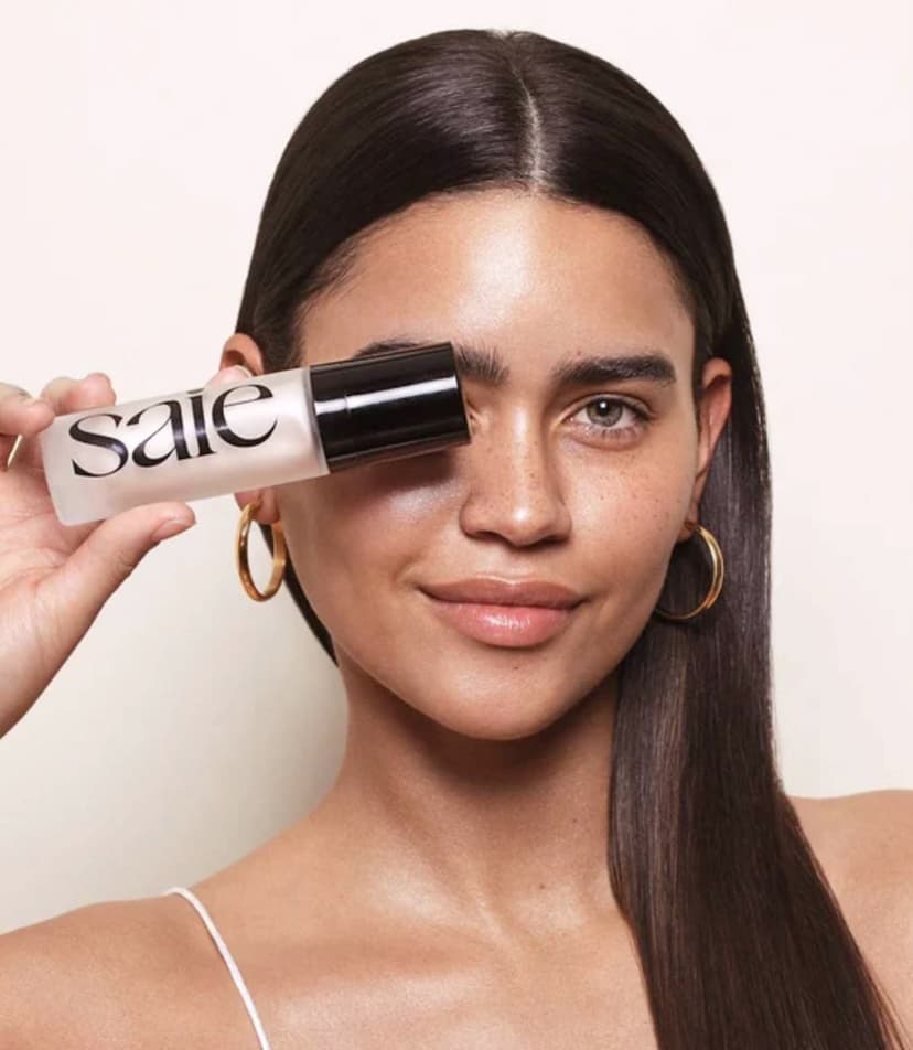 A woman with radiant, freckled, olive-toned skin, dark hair and thick dark eyebrows holds up a package of Saie Beauty's Glowy Super Gel in Starglow. Photo Credit - Saie Beauty