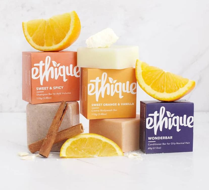 On a marble background, three small, square, paper-wrapped packages from the brand Ethique. The rust-colored package reads Sweet & Spicy Shampoo Bar and has an orange and cinnamon sticks nearby. The orange package has a soap bar on top and reads Sweet Orange Creme Body Wash. The purple box reads Wonderbar Conditioner Bar and has an orange on top. Photo Credit - Ethique