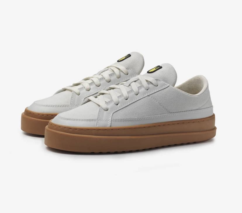 A pair of ivory-white sneakers with white laces, thick rubber soles, and a small black logo on the top of the tongue, made by the brand Unless Collective.