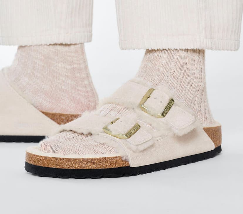 A person's feet shown in cream-colored suede, two-strap sandals with a white fuzzy lining, wearing oat-colored socks underneath and white pants. Photo credit: Birkenstock