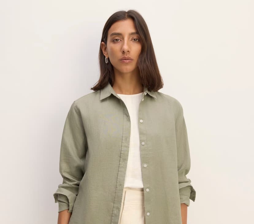 A brunette woman wears a sage green linen blouse, unbuttoned, over a white tee-shirt and cream-colored pants.