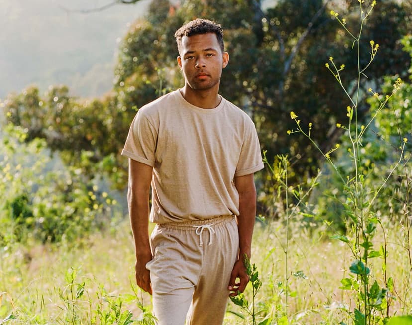 A man wearing tan sweatpants and a matching tan tee shirt stands in a field with trees and yellow flowers.